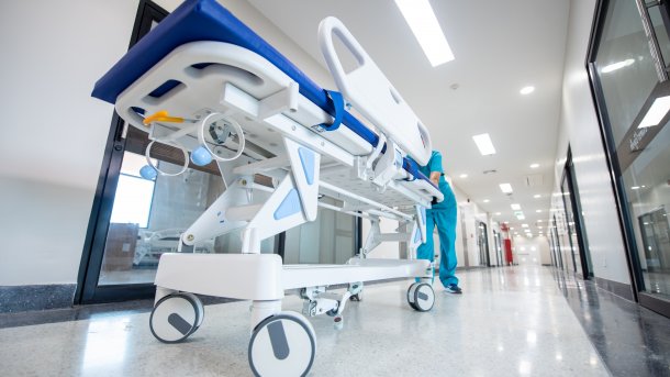 Krankenhausbett wird von Krankenhauspersonal durch einen Krankenhausflur geschoben. Ander Decke ist weißes Krankenhauslicht. Der Boden ist weiß. Alles sieht klinisch aus.
