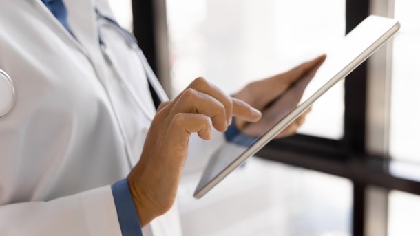 Frau in Weißkittel vor einem Fenster. Ihre eine Hand hält das Tablet, die andere bedient es.