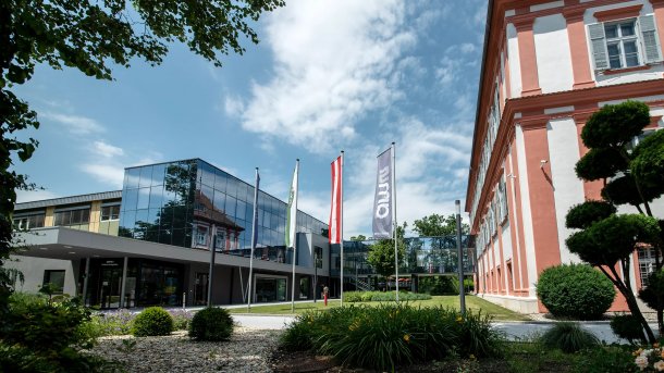 Fahnenstangen mit Flaggen der EU, der Steiermark, Österreichs und dem Firmenlogo ams; rechts davon ein altes Schlossgebäude, links ein modernes Geschäftsgebäude mit Glasfassade