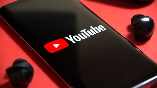 Cell phone with the YouTube logo and lettering on it; black wireless in-ear headphones to the left and right of the phone