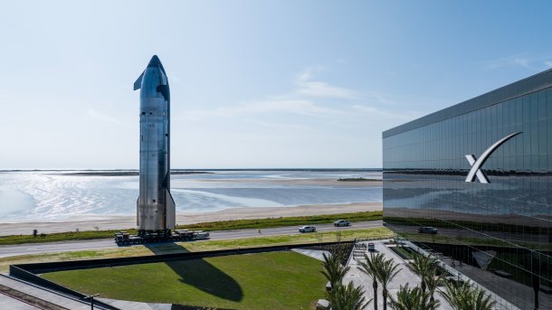 An upright spaceship in front of a SpaceX building