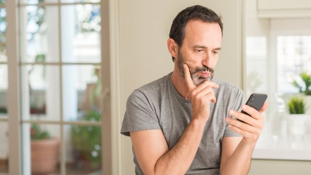 Man in his prime looks at his smartphone and ponders. His right hand is on his chin. He is holding the smartphone in his other hand.