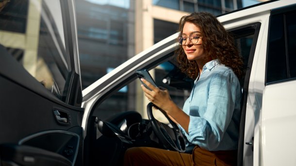 Junge Frau hat ein Smartphone in der Hand und sitzt auf dem Fahrersitz. Eine Hand ist am Lenkrad, die andere hält das Smartphone. Sie schaut auf das Smartphone, ihr Kopf und teilweise auch der Oberkörper sind Richtung Kamera gewandt. Die Fahrertür ist offen. Im Hintergrund ist ein Glasgebäude zu sehen. Die Sonne könnte scheinen, weil das Licht warm ist.