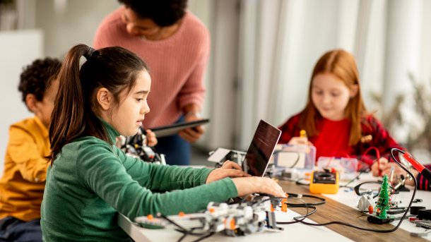 Ein Schulmädchen an einem Laptop, dahinter eine Lehrerin und zwei weitere Kinder