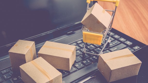 A laptop with a small shopping cart containing a parcel placed on the keyboard. Surrounding it on the keyboard are four more parcels.