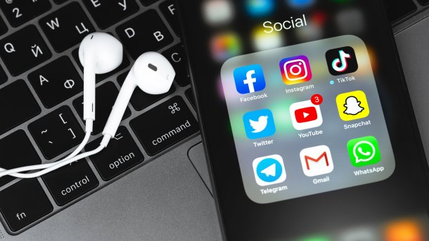 Logos von Social-Media-Apps auf einem Smartphone, das neben Airpods auf einem Macbook liegt