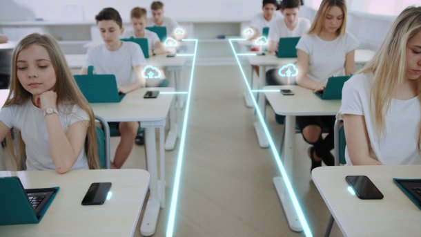 Pupils are sitting in a classroom. A cloud upload sign can be seen on their technical devices