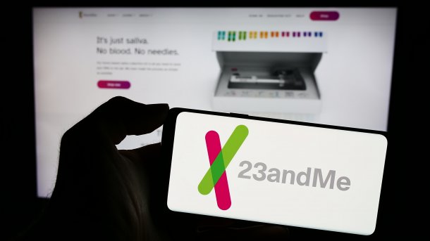 Eine Hand hält ein Handy, dessen Bildschirm "23andMe" zeigt; im Hintergrund unscharf eine Webseite der Firma