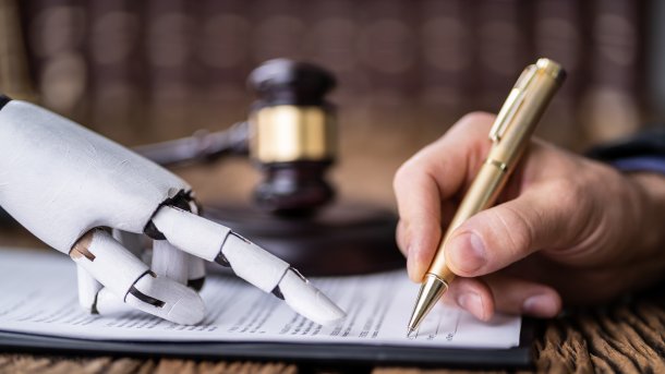 Robot hand points to a piece of paper on which a human hand is writing with a pen. A judge's gavel in the background