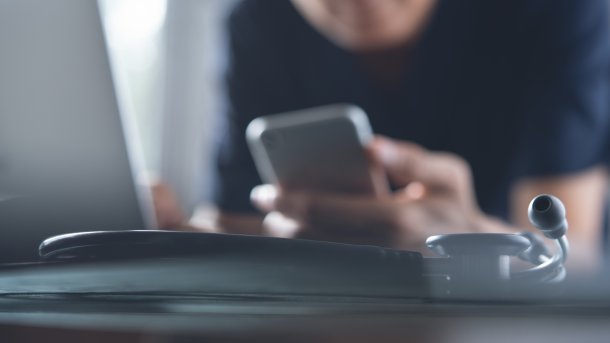 Person nutzt ein Smartphone und sitzt vor einem Computer. Daneben ist ein Stethoskop.