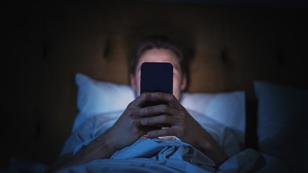 Man in bed holding smartphone in his hand, covering his face