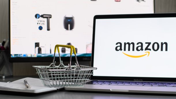 Miniature shopping basket to the left of a laptop with the Amazon logo. In the background are Amazon Buy Boxes, where products are placed on the website of online giant Amazon.