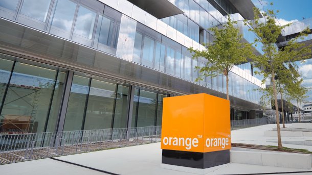 Orange Würfel mit weißer Aufschrift "orange" steht im Hof eines Bürogebäudes mit viel Glas