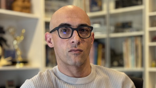 A man with black smart glasses sits in front of a bookshelf.