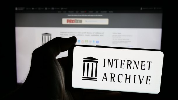 Das Logo des Internet Archive wird auf einem Smartphone angezeigt. Im Hintergrund ist auf einem Monitor die Startseite mit dem Logo der Wayback machine zu sehen.