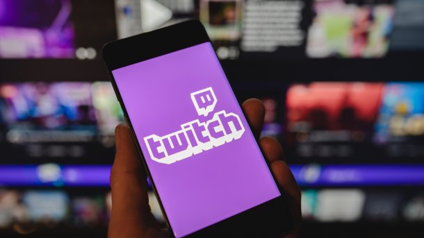 Smartphone mit dem Twitch-Logo auf dem Bildschirm vor einem verschwommenen Hintergrund mit mehreren Monitoren.