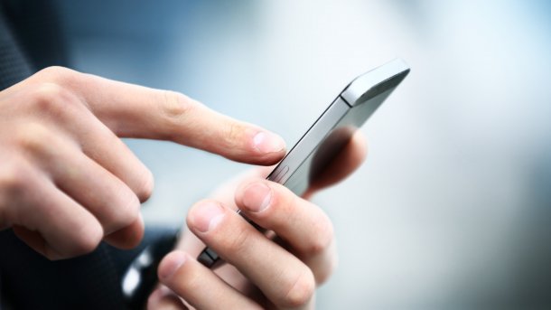 Eine Hand hält ein Smartphone, der Zeigefinger der anderen Hand tippt auf dessen Bildschirm