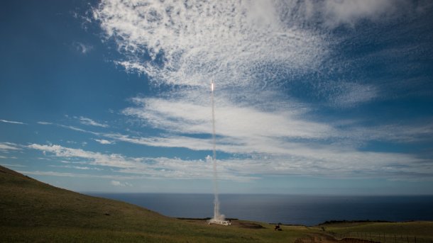 Rocket taking off from a green meadow, the ocean in the background.