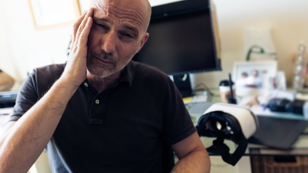 Man at a desk holds his hand to his head.  VR glasses are lying on the desk.
