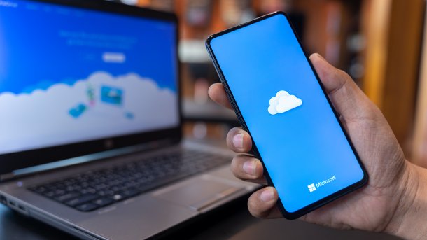 Offener Laptop, davor hält eine Hand ein Smartphone; beide Bildschirme zeigen symbolisch die Microsoft-Cloud