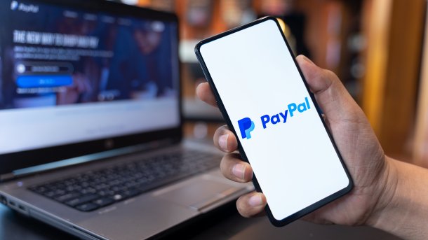 A hand holds a smartphone up to the camera; the Paypal logo can be seen on the display