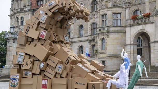 Many stacks of paper as a sign against excessive bureaucracy. Three figures in hospital uniforms bend down in front of the pile. 