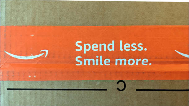 Close-up of an Amazon parcel. The orange adhesive tape next to the Amazon logo reads "Spend less. Smile more."