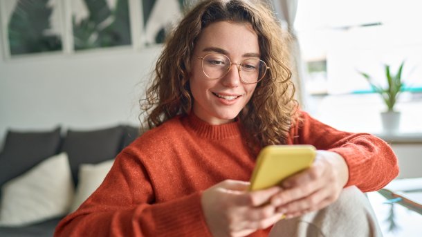 Young woman with reddish-blonde curls and orange sweater on her smartphone in a living room.