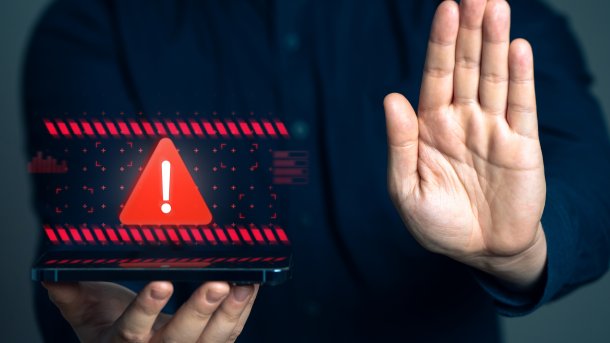 Eine Person hält ein mobiles Endgerät, über das ein Achtung-Zeichen retouchiert ist, in ihrer rechten Hand, während sie ihre linke Hand abweisend in die Kamera hält.