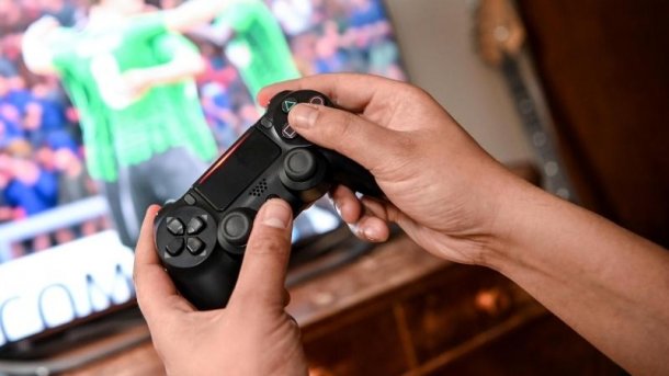 Young gamer wearing a hoodie and headphones in front of a computer screen. He is playing a video game in a darkened room with purple lighting.