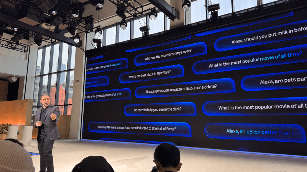 On the back wall of a stage are numerous questions that users can ask Alexa+, including "Who has the most Grammys ever?" or "What is the most popular movie of all time?"; at the front of the stage, a man in a dark grey robe and trainers gestures.