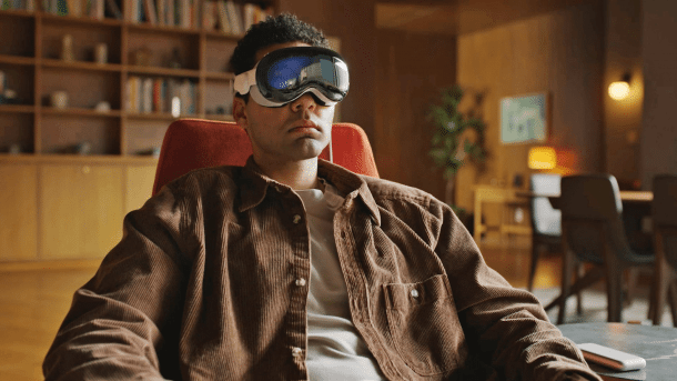 A man sits on a red armchair in a living room, motionlessly wearing VR goggles.
