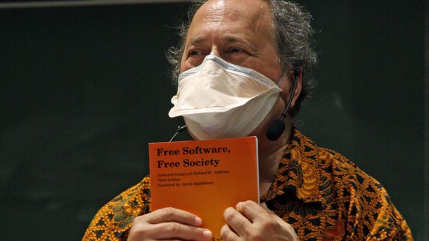 Richard Stallman, wearing an antiviral mask over his mouth and nose, holds up his book titled "Free Software, Free Society" during his appearance at TU Munich.