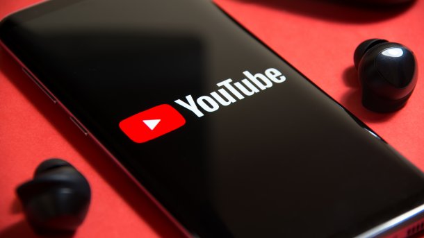 Mobile phone showing the YouTube logo and lettering; a black, wireless in-ear headphone lies to the left and right of it.