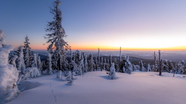 Im makellosen Schnee führen Spuren durch in eine endlos stille Winterlandschaft. Die klare Luft lässt die Farben des Sonnenuntergangs besonders pastellig erscheinen. Das Licht malt sanfte Schatten und schafft so eine Tiefe ohne Härte