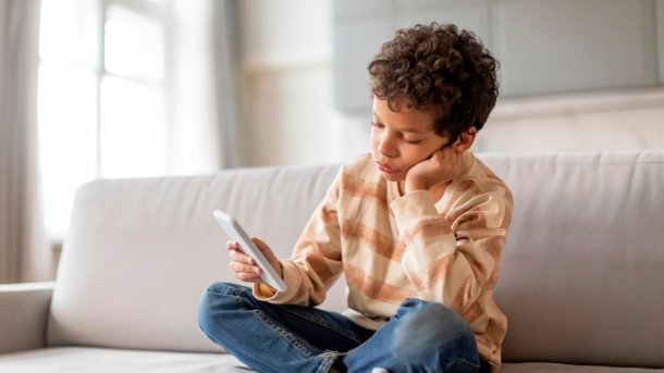 Gelangweiltes Kind mit einem Smartphone in der Hand, das auf einem Sofa sitzt