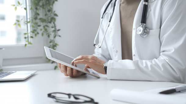 Mann im Weißkittel am Tablet, vor ihm ein Laptop.