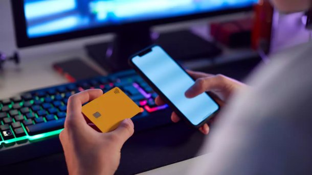 Person holding a credit card in one hand and a smartphone in the other. A computer monitor is in the background.