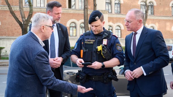 Ein Polizist in Uniform, flankiert von zwei Herren in Anzügen, scannt mit seinem Handy die Hand eines dritten Herren in Anzug; im Hintergrund die rosafarbene Wand der Rossauer Kaserne in Wien-Alsergrund.