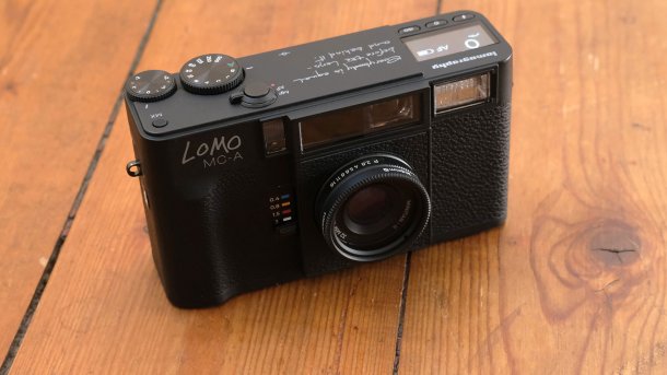 Schwarze analoge Kompaktkamera Lomo MC‑A mit festem Objektiv auf einem Holztisch fotografiert.
