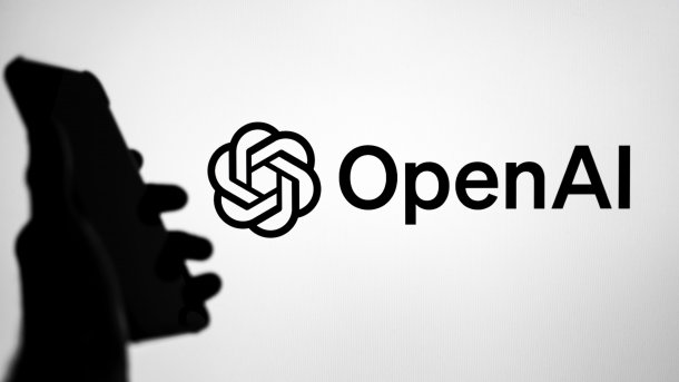 OpenAI-Logo auf Bildschirm, davor Hand mit Smartphone in Silhouette