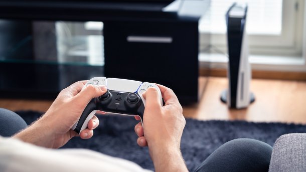 Mann hält Playstation-Controller, im Hintergrund Playstation 5