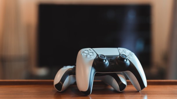 Zwei Playstation-Controller auf einem Holztisch, im Hintergrund ein verwaschener Fernseher