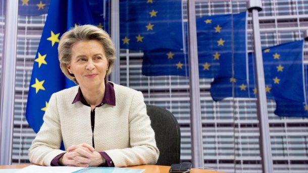 Ursula von der Leyen sits at a table, with flagpoles bearing EU flags visible behind her