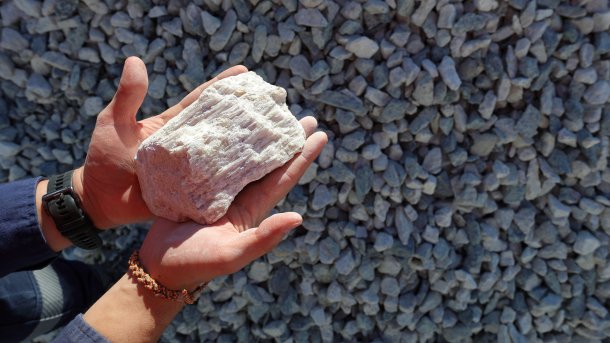 A hand holds a chunk of the lithium-containing mineral spodumene. Stones are visible in the background.