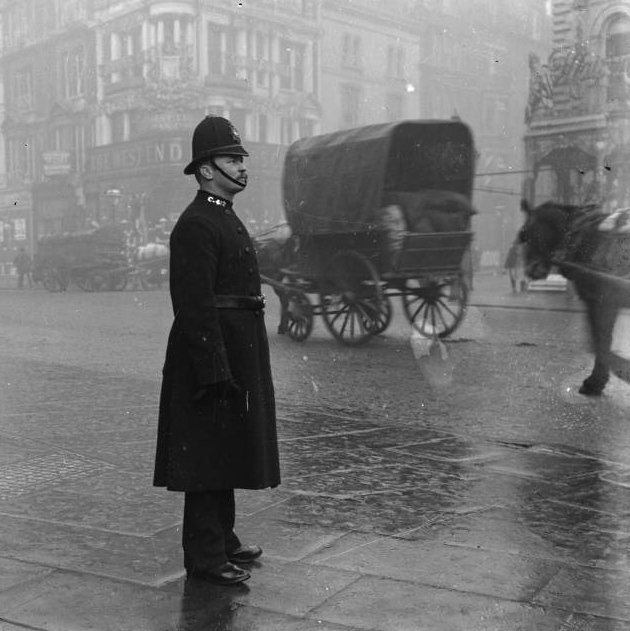 Altes Foto eines Polizisten im Regen, dahinter Pferdefuhrwerke