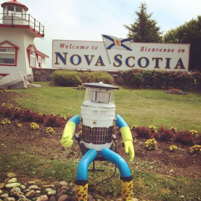 Der hitchBOT vor einem Schild &quot;Welcome to Nova Scotia&quot;
