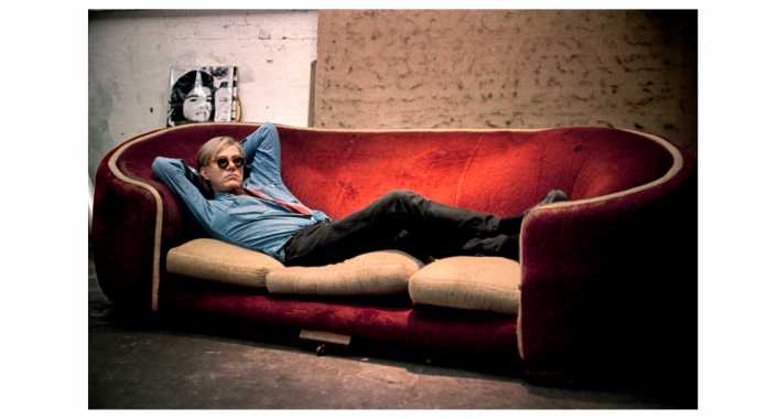 Andy Warhol at 47th street Factory,  poses on notorious Factory red couch with Jackie Kennedy silkscreen.1965 New York City