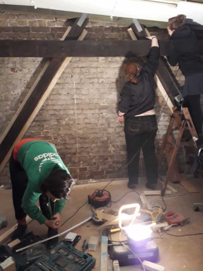 Drei Leute halten Werkzeuge und Holzbalken in einem unverputzten Raum