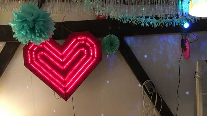 Ein großes leuchtendes rotes Herz hängt an einer weißen Wand mit Partydekoration